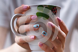 Girlish manicure with volumetric ladybugs on pink and blue fingernails. Female hands holding a cup with lady-bird and clover leaf