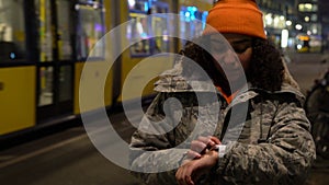 Girll young woman using smart watch at night with trams by Alexanderplatz Station, Berlin, Germany