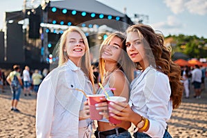 Girls cheer colorful drinks at beach music fest. Happy friends enjoy sunny day, summer party vibes. Dance, laughter near