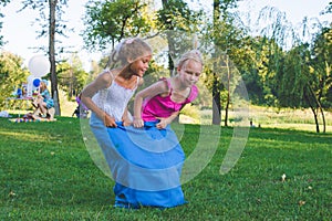 Girls compete in a relay race . Jumping in bags . They laugh and fall .