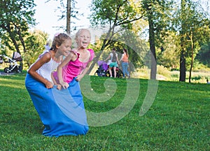 girls compete in a relay race . Jumping in bags . They laugh and fall .