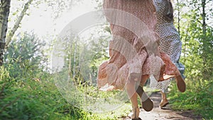 Girls in light dresses are running in a sunny forest. Girls` legs in close-up. The girls hold hands and run down the path