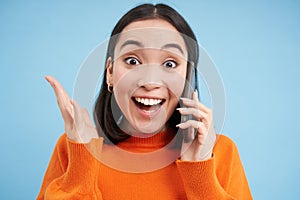 Girls reaction to amazing news she received on mobile phone. Happy asian woman looks surprised and excited, blue