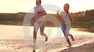 Girls run on the sand along the shore on the beach splashing water drops and laughing. Happy and free teenagers on a