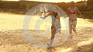 Girls run on the sand along the shore on the beach splashing water drops and laughing. Happy and free teenagers on a
