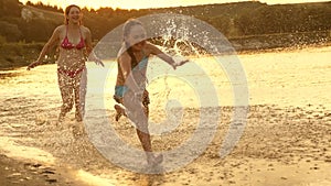 Girls run on the sand along the shore on the beach splashing water drops and laughing. Happy and free teenagers on a