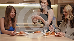 Girls sit and eat at the table in a modern kitchen in an apartment. The landlady comes with three tall and thin glasses