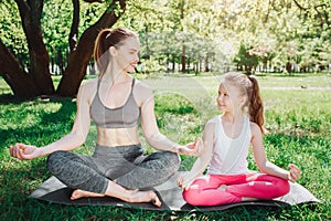 Girls are sitting on carimate in lotus pose and loking at each other. They are smiling. Girls are excercissin outside in