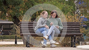 Girls students sitting in park on bench with a modern digital tablet. Teens from university, college, school, teach