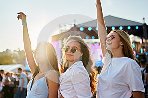 Girls in sunglasses at sunset, arms raised, dancing on sandy beach with festival stage background. Female friends enjoy