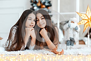 Girls twins in front of the fir-tree. New year`s eve. Christmas. Cozy holiday at the fir-tree with lights