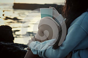 A girlsitting on sea rock, holding a cup of morning tea or coffee in the beach. Self care, relax weekend concept.