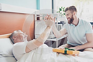 Give me five! Young stylish red bearded guy is visiting and cheering up his grandpa lying in bed at hospital ward