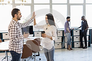 Give me high-five. Two coworkers giving high-five and holding coffee cups with smile while sitting on the couch at office with