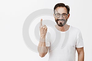 Give me one minute to dress up. Charming carefree friendly-looking bearded man in trendy glasses, showing index finger