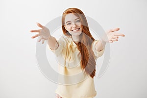 Give me warm hug. Studio shot of friendly cute redhead female student pulling hands towards camera, tilting head and