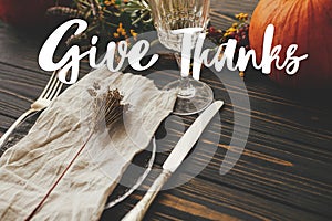 Give thanks text on plate, cutlery,  napkin on wooden table with pumpkins and autumn flowers, dinner rustic setting. Happy