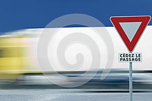 Give way yield french cÃ©dez le passage road sign, motion blurred truck vehicle traffic background, white signage triangle red