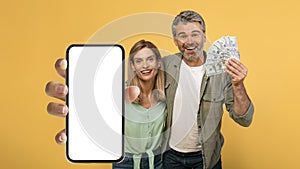 Giveaway concept. Emotional middle aged couple holding lot of dollar cash, showing smartphone with blank screen, mockup