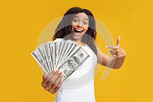 Giveaway concept. Excited young black woman holding a lot of dollar cash, showing big win over yellow studio background