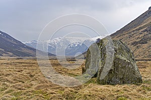 A Glacial erratic stands tall in the Valley floor at Glen Esk in the Angus Glens, with snow covered mountains in the distance.