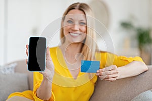 Glad attractive young blonde caucasian woman in yellow clothes shows smartphone with blank screen and credit card