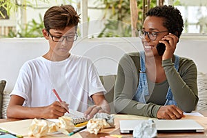 Glad black girl with short curly hair, has telephone conversation, looks positively at her groupmate who writes down