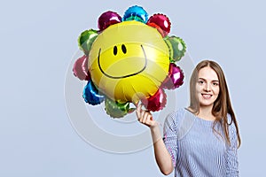 Glad brunette woman wears striped blouse, holds balloon, happy to celebrate daughter`s birthday, meets guests, isolated over blue