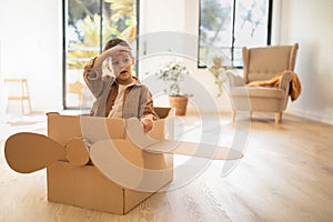 Glad caucasian little child capitan sitting in cardboard box airplane, has fun alone in room interior