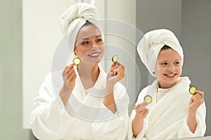 Glad caucasian small girl and young lady in bathrobe and towel, have fun with pieces of cucumber enjoy beauty care