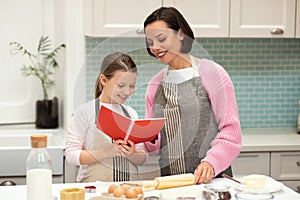 Glad caucasian young mom and small daughter in aprons make cookie dough, look at recipe book in kitchen