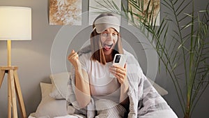 Glad Caucasian young woman holding mobile phone clenched fist gets message with excellent news wrapped in soft blanket enjoys good