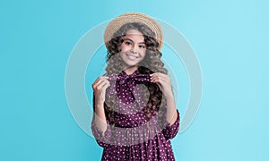 glad child in straw hat with long brunette curly hair on blue background