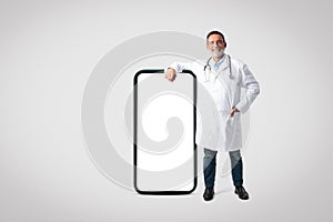 Glad confident european senior doctor in white coat with stethoscope near big screen phone