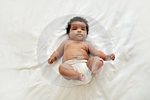 Glad cute african american little curly baby in diaper lies on white soft white bed, waking after sleep