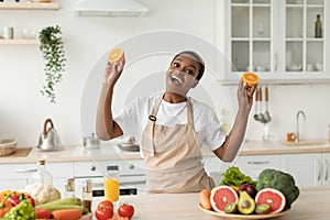 Glad emotional young black woman in apron have fun on minimalist kitchen interior, holds oranges