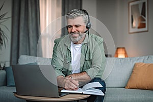 Glad european aged man retired with beard in headphones looks at laptop, work in room interior