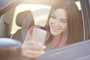 Glad European female with long dark hair, happy to recieve message on smart phone, poses in car, stops on road, connected to wirel