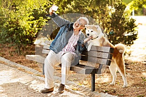 Glad european old man in casual enjoys walking with dog, sit on bench in park, taking photo on smartphone
