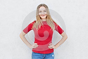 Glad female with light hair and blue eyes having smile on her face wearing red sweater and jeans holding hands on waist. Attracriv