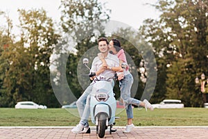 Glad girl in trendy jeans kissing boyfriend on scooter on blur nature background. Caucasian man in white clothes
