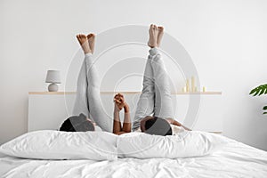 Glad happy young african american husband and wife in domestic clothes are lying on bed, holding hands with raised legs