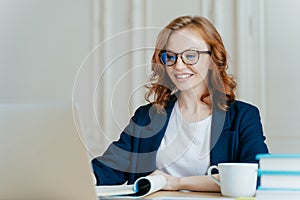 Glad lovely woman has ginger hair, positive smile, sits with laptop computer at desktop, happy to make video call or conference,