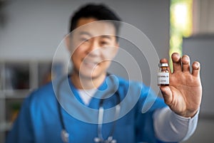 Glad mature chinese man doctor show vaccine in clinic office interior, blurred