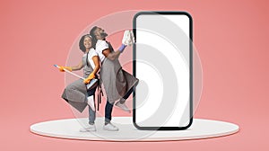 Glad millennial black couple in aprons have fun, play at virtual guitar, enjoy cleaning near huge phone