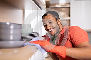 Glad millennial black man in rubber gloves wipes dust from shelf with dishes enjoy purely in white kitchen photo