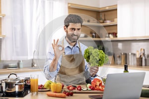 Glad millennial caucasian male cooking eat show salad and ok hand sign at table with vegetables and computer