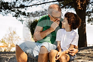 Glad old caucasian man hugs little mixed race kid, whispers secret in ear, enjoy spare time outdoor