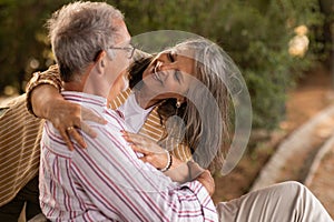 Glad senior caucasian woman hugging husband, sitting on bench, have fun in park