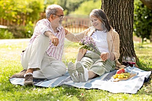 Glad senior european man gives bouquet to woman, enjoy romantic date, picnic and lunch in park, outdoor